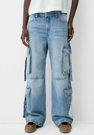 Person iført lyseblå vidde denim cargobukser med store sidelommer og brune mønstrede sko, stående mod en hvid baggrund.