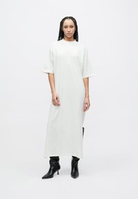 Lang, løstsittende hvit kjole laget av mykt stoff, med korte ermer og en rett silhuett. Kombinert med svarte støvletter med hæl.