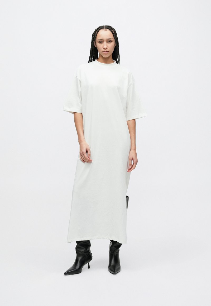 Lang, løstsittende hvit kjole laget av mykt stoff, med korte ermer og en rett silhuett. Kombinert med svarte støvletter med hæl.