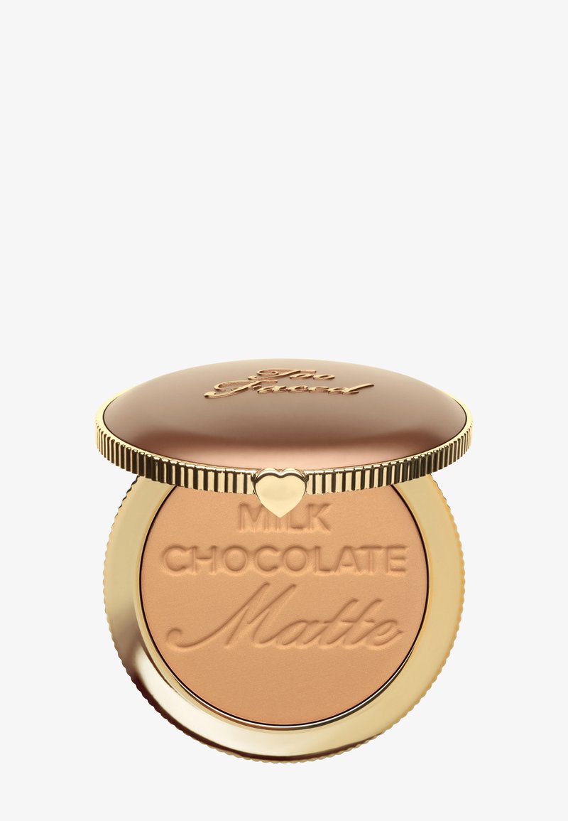 Maquillage en poudre compacte bronze et or étiqueté « Milk Chocolate Matte » avec un fermoir en forme de cœur sur le couvercle, partiellement ouvert sur un fond blanc.