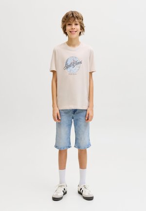Ragazzo sorridente in piedi, indossa una t-shirt beige chiaro con grafica, pantaloncini di jeans blu sbiaditi, calzini bianchi e scarpe da ginnastica bianche con strisce nere.