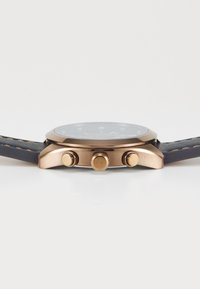 Gros plan sur le côté d'une montre-bracelet avec boîtier en or rose, deux boutons, couronne, et bracelet en cuir noir avec coutures beiges sur fond blanc.