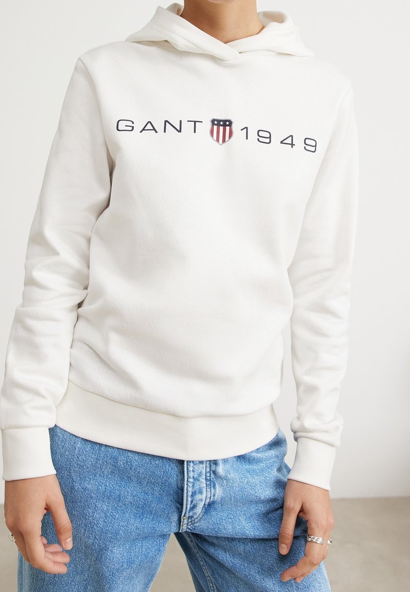 Personne portant un sweat à capuche blanc GANT avec logo et texte 1949, associé à un jean bleu clair, debout avec les mains détendues le long du corps.