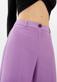 Fialové kalhoty s širokými nohavicemi vyrobené z měkké tkaniny, s předním zapínáním na knoflík, bočními kapsami a jemnými růžovými švy.