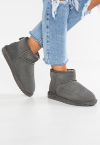Graue Wildleder-Stiefeletten mit einer weichen Textur, abgerundeter Zehenpartie und einem niedrigen Absatz. Das Design umfasst einen umklappbaren Schaft und einfache Nähdetails.