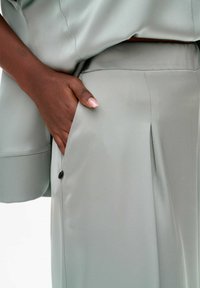Lichtgrijze stoffen broeken met een geplooid ontwerp, voorzien van een zijzak en een knoopaccent. Gladde textuur, slim fit.