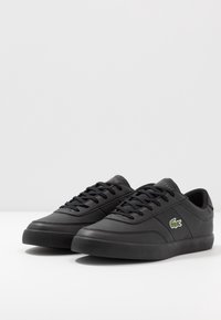 Paire de baskets basses noires avec lacets noirs, panneaux latéraux texturés et un petit logo crocodile vert sur les côtés extérieurs.
