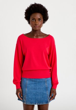 Kvinde med mørkt krøllet hår iført gyldne hoop-øreringe, en rød langærmet sweater og en blå denimnederdel, stående mod en ensfarvet hvid baggrund.