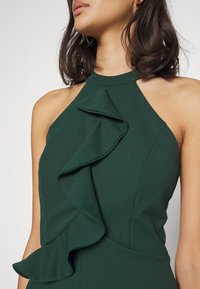 Mörkgrön klänning med halterneck-design, med en framträdande volangdetalj framtill, slät textur och åtsittande silhuett.