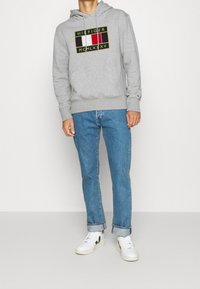 Muž v šedé mikině Tommy Hilfiger, modrých zrolovaných džínách a bílých teniskách Veja, stojící na pozadí s jemným světlem.