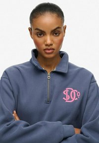 Kobieta z spiętymi włosami, ubrana w niebieską bluzę z zamkiem częściowym, z różowym haftowanym logo, stojąca na jednolitym białym tle.