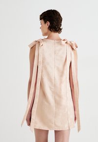Frau von hinten zu sehen, die ein hellrosa ärmelloses Kleid mit floralem Muster und langen gebundenen Schleifen an den Schultern trägt.