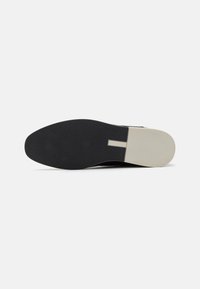 Suela negra de zapato con una parte inferior de goma negra texturizada y una sección de punta beige en contraste. Presenta un corte rectangular para mayor estilo.
