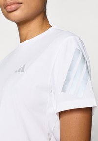 Fehér sport póló rövid ujjakkal, texturált anyaggal és fényvisszaverő csíkozással a bal vállon.