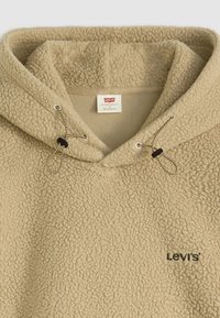Béžová sherpa fleece mikina s kapucňou s nastaviteľnými šnúrkami a vyšitým logom Levi's na hrudi, so štítkom so športovým strihom vo vnútri.
