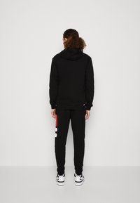 Felpa nera con cappuccio regolabile, abbinata a pantaloni tuta neri con un logo rosso e bianco, indossata con sneakers bianche.