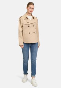 Beige kaksirivinen trench-coat suurilla nappilla, olkapäälenkkien ja sivutaskujen kanssa, yhdistettynä istuviin sinisiin farkkuihin ja valkoisiin lenkkareihin.