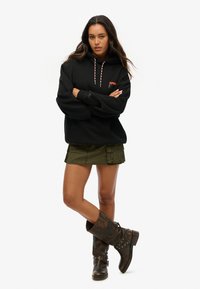 Hoodie noir surdimensionné avec des cordons contrastés rouges et blancs, associé à une mini jupe cargo verte et des bottes marron hautes jusqu'aux genoux.