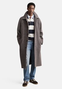 Pilkas houndstooth paltas su plačiais atlapais, užsegamas sagomis, su šoninėmis kišenėmis, dėvimas ant tamsiai mėlynos dryžuotos marškinių ir mėlynų džinsų.