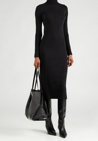 Frau in einem schwarzen langärmligen Rollkragenkleid mit schwarzen knielangen Stiefeln, die eine große schwarze Leder-Handtasche hält.