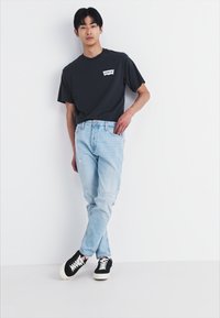 Donkergrijze t-shirt met een wit logo, lichtblauwe versleten jeans en zwarte sneakers met witte zolen. Casual pasvorm met een ontspannen ontwerp.
