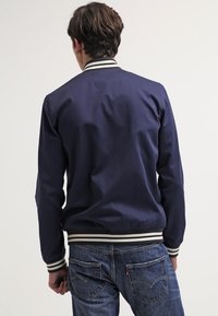 Veste aviateur bleu marine avec col et poignets côtelés rayés, présentant une texture de tissu lisse. Portée avec un jean en denim bleu.