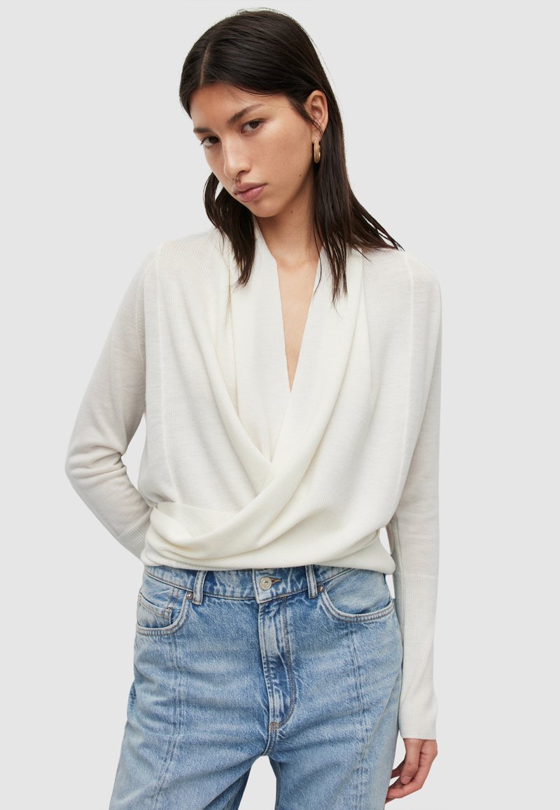 Weißes, langärmliges Oberteil mit tiefem V-Ausschnitt, aus weichem Stoff gefertigt. Kombiniert mit hellblauen, hochgeschnittenen Jeans mit einem ausgewaschenen Muster.