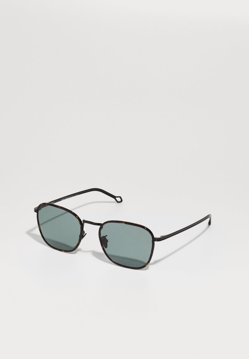 Ochelari de soare cu ramă cu model tartan, lentile verde închis, accente metalice și brațe subțiri negre, de formă pătrată.