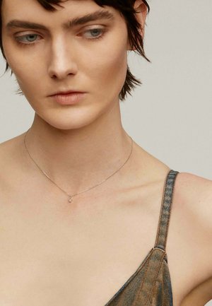 Collar de plata con un pequeño colgante en forma de lágrima. Llevado con una prenda de denim que cuenta con una tirante delgado. Tono de piel clara y cabello corto visible.