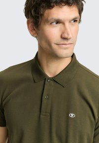 Olivgrünes Poloshirt aus strukturiertem Stoff mit klassischem Kragen, zwei Knöpfen und kleinem Logo auf der linken Brust.