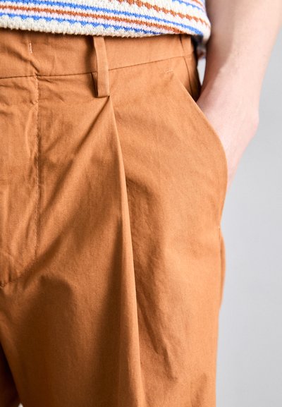 Pantalons en coton marron avec des plis, poches latérales et une texture lisse, accompagnés d'un pull rayé multicolore à la taille.