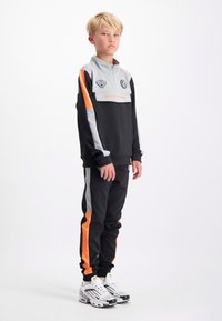 Zwarte en grijze trainingsoutfit met oranje accenten en een kwart-rits. Bevat logo-patches en zijstrepen. Draagt met witte sportschoenen.