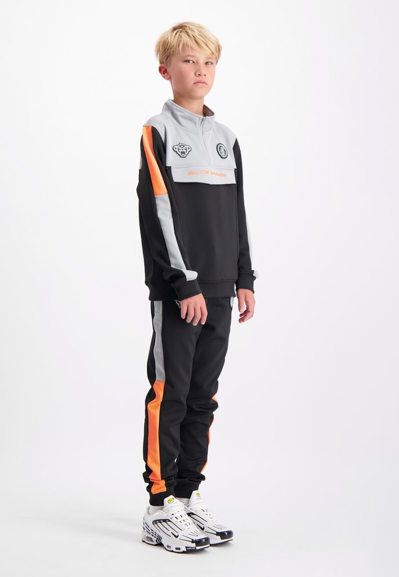 Zwarte en grijze trainingsoutfit met oranje accenten en een kwart-rits. Bevat logo-patches en zijstrepen. Draagt met witte sportschoenen.
