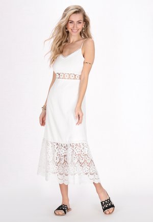 Witte midi-jurk met kantafwerking, spaghettibandjes en decoratieve uitsnijdingen in de bloemenriem. Gecombineerd met zwarte slidesandalen.