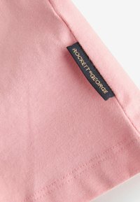 Esquina de tela rosa con una etiqueta rectangular negra cosida en el borde que dice "ROCKETT St GEORGE" en letras doradas.