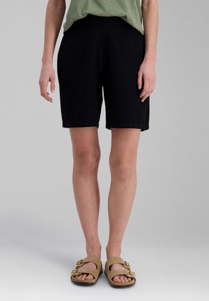 Person trägt schwarze kniehohe Shorts, grünes Oberteil und beige Sandalen mit doppelter goldener Schnalle und steht vor grauem Hintergrund.
