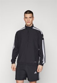 Zwarte sportjack met witte zijstrepen, volledige rits, hoge kraag, elastische manchetten en tailleband, gecombineerd met zwarte shorts met logo-accent.