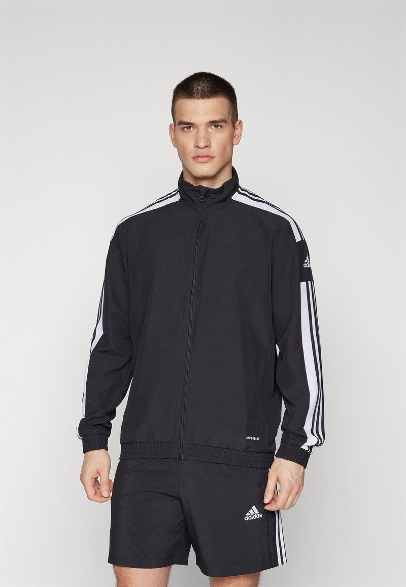 Zwarte sportjack met witte zijstrepen, volledige rits, hoge kraag, elastische manchetten en tailleband, gecombineerd met zwarte shorts met logo-accent.