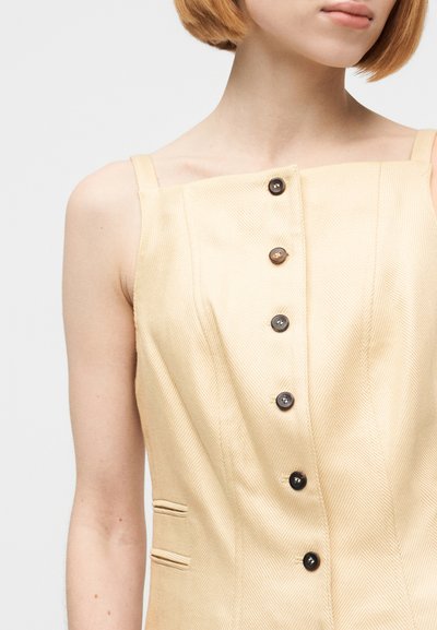 Γυναίκα με κοντά κόκκινα μαλλιά, φοράει ένα μπεζ αμάνικο φόρεμα με κατακόρυφα κουμπιά και δύο οριζόντιες σχισμές-τσέπες στο πλάι.