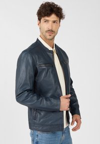 Marineblaue Lederjacke mit Stehkragen, Front-Reißverschlusstasche und dezenten Schulternähten, kombiniert mit einem leichten Hemd darunter.