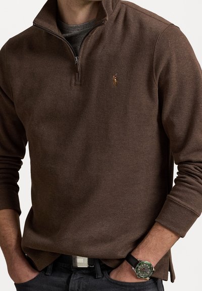 Pull à fermeture éclair marron en tissu tricoté, avec un petit logo sur la poitrine. Associé à une montre et un pantalon noir.