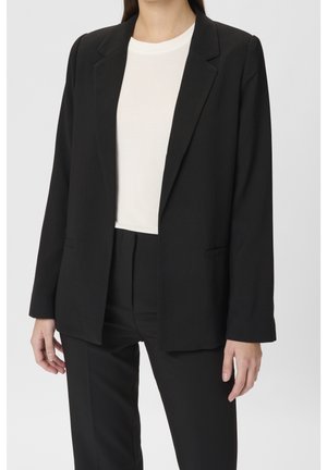 Femme portant un blazer noir sur mesure et un pantalon assorti, avec un haut blanc à col rond, posant devant un fond gris uni.
