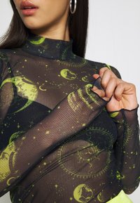 Pitkähihaisessa mustassa kangaspuserossa on vihreitä taivaankappaleita, ja siinä on korkeakauluksellinen muotoilu sekä teksturoitu ribbikangas. Tiukka istuvuus korostaa muotoa.