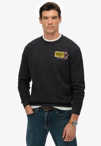 Sudadera negra con cuello redondo, que presenta un parche de logo amarillo y rojo. Llevada sobre una camisa blanca, combinada con jeans azules.