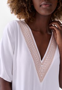 Witte top met een V-hals met ingewikkelde beige kantdetails. Korte mouwen en zachte textuur, die het halslijnpatroon accentueert.