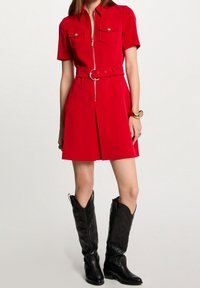 Rode, korte mouw jurk met een ritssluiting aan de voorkant, twee borstzakken, een ceintuur in de taille en een geplooide rok. Draagt met zwarte cowboylaarzen.