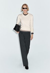Maglione a V crema, pantaloni a gamba larga grigio scuro e borsa nera. La modella indossa occhiali da sole. Texture lisce e design minimalista.