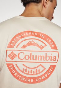 Svetlá krémová polokošeľa s krátkym rukávom, ktorá obsahuje veľký koralový grafický motív s horami, stromami a textom "Columbia Sportswear Company."