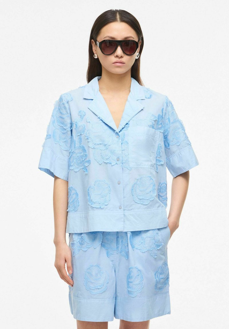 Femme portant une chemise bleu clair à manches courtes boutonnée et un short assorti avec des motifs floraux brodés, accessoirisée de grandes lunettes de soleil foncées.