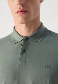 Grønn polo skjorte med teksturert stoff, som har en tre-knapps placket, grå striper på kragen, og en tonet "BOSS" logo på brystet.